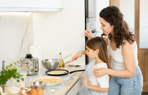 Obraz na plátně Mom and daughter are cooking in kitchen, frying pancakes