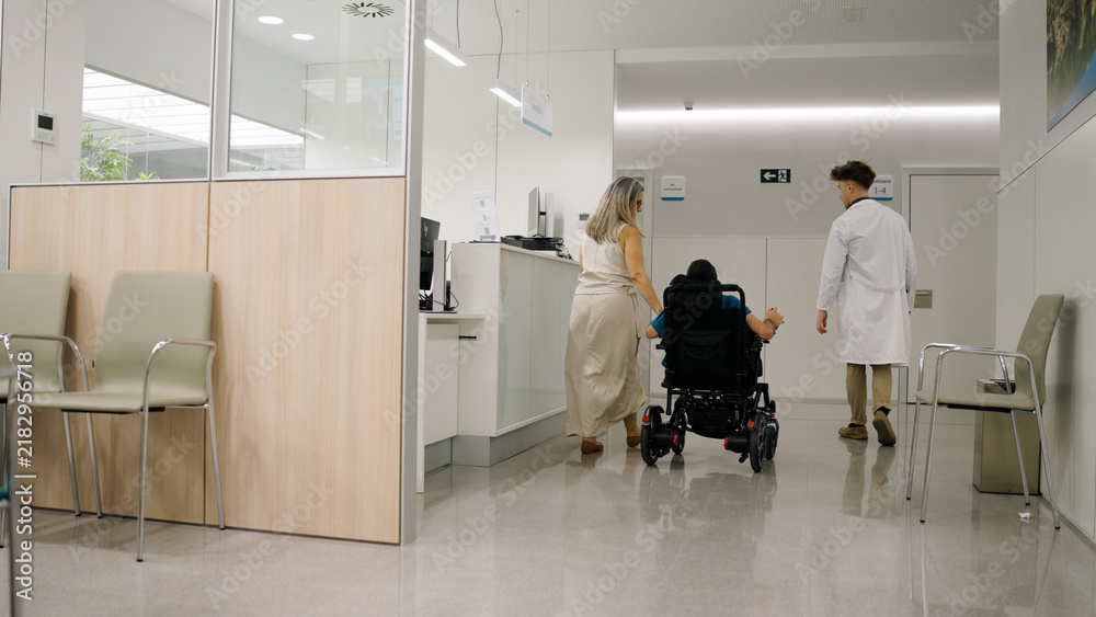 Fototapeta premium Woman pushing a person in a wheelchair with a doctor walking beside them in a modern hospital corridor, showing healthcare and support