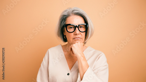 Senior woman with grey hair wearing glasses, looking pensive and serious with her hand on her chin, thinking and reflecting