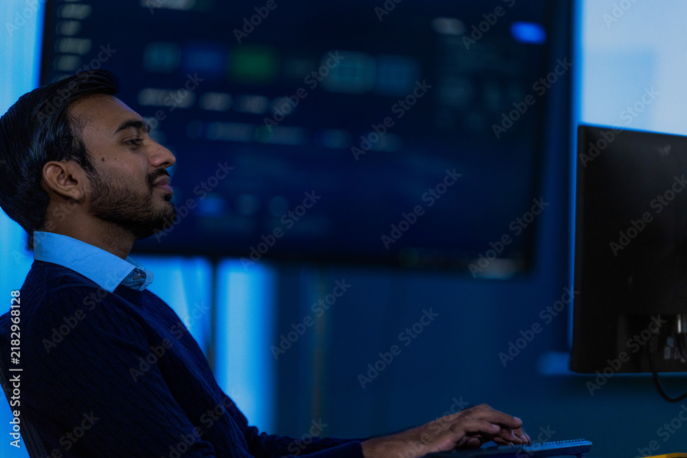 Fototapeta premium A male data analyst in a dark navy suit jacket and light blue shirt watches a chart update while deciding whether to escalate in a blue lit monitoring room in a modern technology company