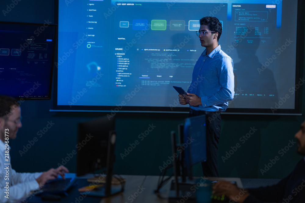 Fototapeta premium A young male analyst with dark curly hair and clear framed glasses in a light blue shirt raises an open palm to emphasise a point about network security in a blue lit monitoring room