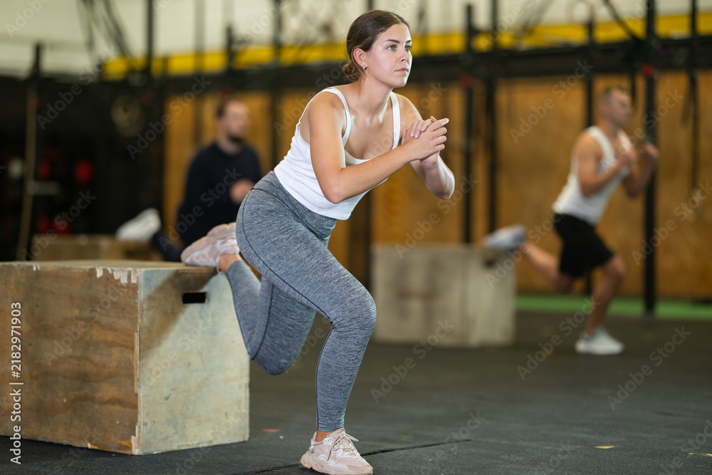 Fototapeta premium Concentrated young girl in fitness attire performing Bulgarian split squats using wooden plyometric box at gym. Bodyweight training concept