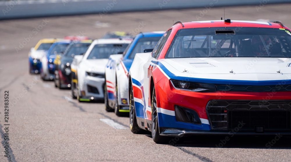 Fototapeta premium Racing cars lined up on pit lane of speedway track. Red white and blue stock race car in foreground with row of competitors behind. Motorsport competition and auto racing speed concept.