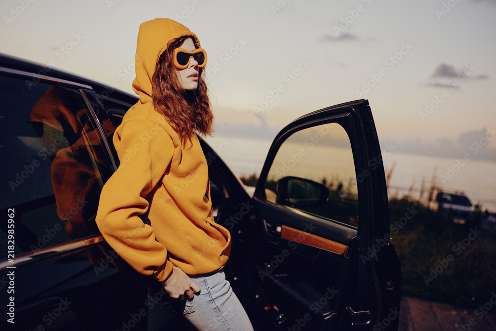 Obraz premium Person in a yellow hoodie and sunglasses stands beside an open car door at sunset, capturing casual fashion and street style in an outdoor portrait.