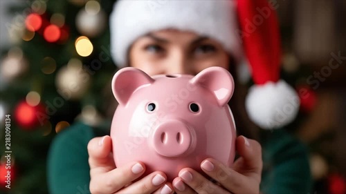 Woman in santa hat holding pink piggy bank near christmas tree. Christmas savings and holiday budget concept. Woman saving money for new year celebration