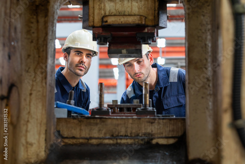 Naklejka Two Caucasian male engineers seen through machine frame inspecting press die in factory