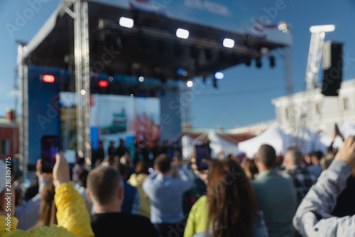 Back view of an audience watching live concert in blur or public event on outdoor stage setup on sunny day, with people holding up smartphones to capture show