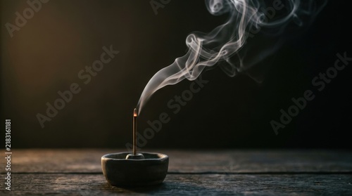 Incense stick burning in a small ceramic holder emitting delicate white smoke on a wooden surface.
