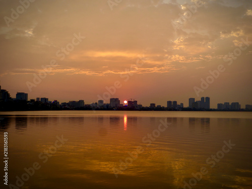 Golden Sunset Over Tranquil Urban Lake with City Skyline and Glowing Sun Reflection on Calm Water on West Lake in Hanoi, Vietnam