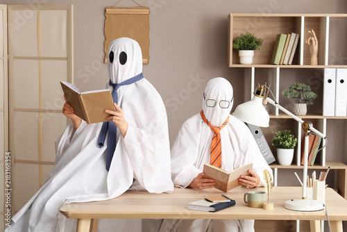 Two people dressed as ghosts with ties reading books at an office desk  halloween and business