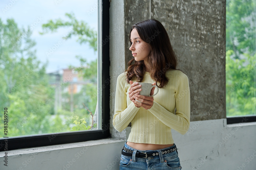 Obraz premium Pensive young woman holding mug standing near window and looking outside