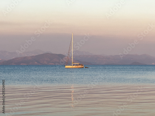 Anchored sailboat on calm Garitsa Bay at pastel sunset, Corfu Town, Greece, hazy horizon