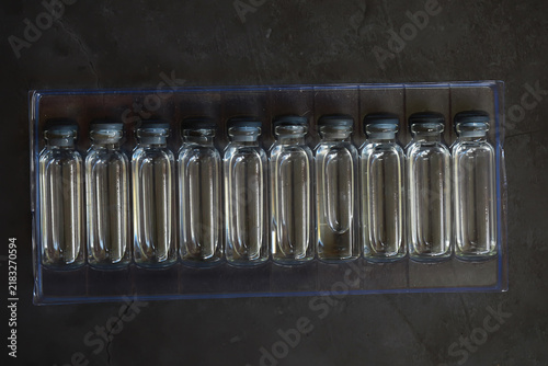 Принти на полотні Row of Clear Glass Bottles with Black Caps on Transparent Tray Against Dark Back