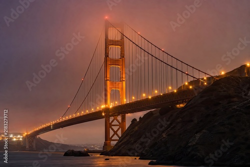 Golden gate bridge in san francisco california during purple sunset illuminates orange lights reflecting on water with misty fog for travel and landmark tourism.