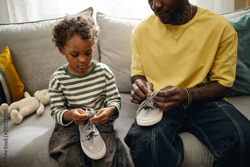African American father helping male child tying shoelaces indoors. Concept depicts early childhood development, ideal for parenting education, family lifestyle marketing