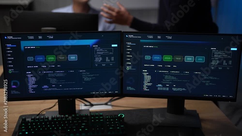 Twin monitors on a shared desk glow with an AI agent pipeline builder and a coloured node graph as blurred teammates move behind them inside a dark AI startup office.