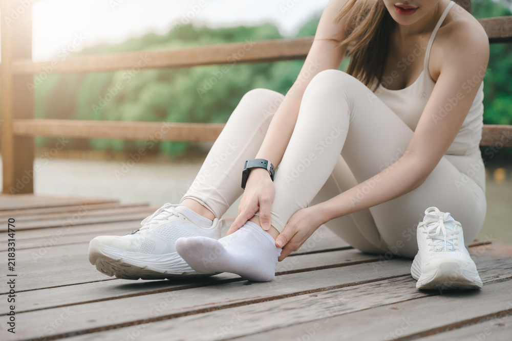 Fototapeta premium Fitness woman runner feeling pain on ankle and leg during exercise sitting on wooden bridge outdoors, holding injured foot with hands after sprain or muscle strain injury