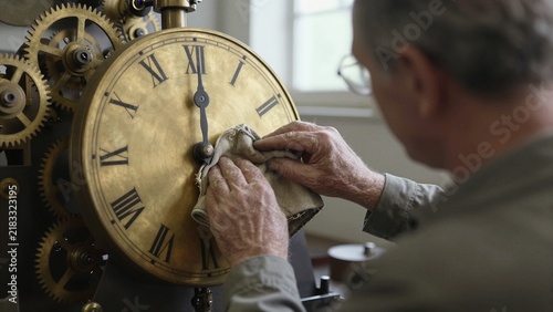 An elderly clockmaker meticulously cleans a vintage brass timepiece with Roman numerals and exposed gears in a softly lit workshop.