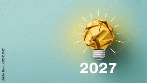 A glowing yellow crumpled paper light bulb representing a creative idea for the year 2027 against a solid light blue background, studio shot