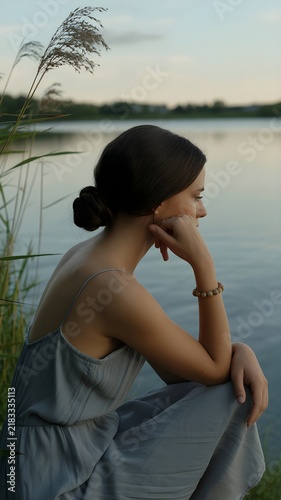 Mujer pensativa en vestido gris contemplando el lago en calma al atardecer