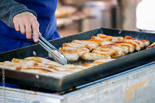 Cuadro en lienzo Turning Sizzling Bratwurst Sausages with Tongs on an Outdoor Flat Top Grill