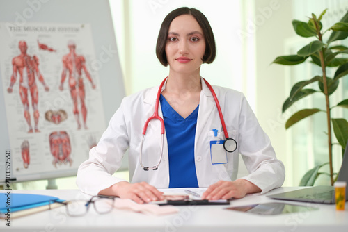 Young physician in blue scrubs and white coat sits at desk in modern clinic, looking at camera with steady expression. Medical poster, laptop, files, and indoor plant create clean healthcare setting