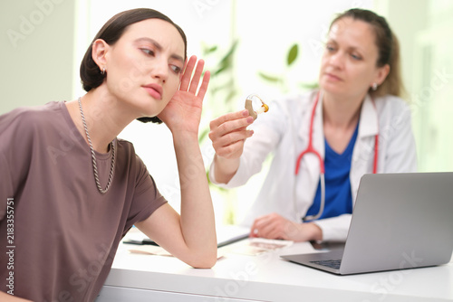 Young woman cups ear while audiologist explains hearing aid during consultation in bright medical office, with laptop on desk, soft daylight, focused expression, hearing care treatment, and patient