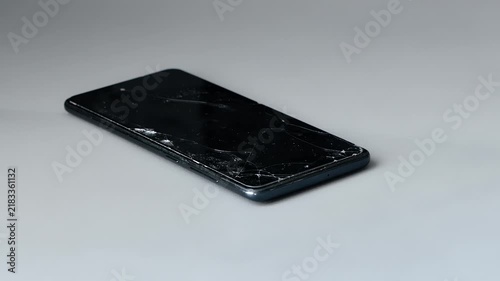 Cracked screen protector for smartphone. Damaged mobile phone with shattered front glass laying on plain grey studio background.