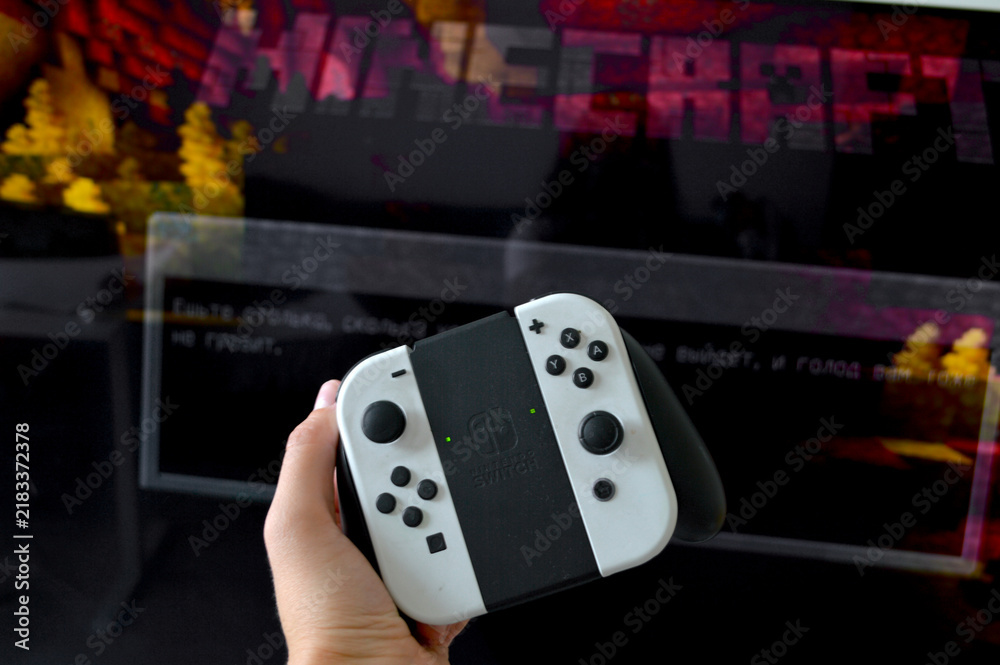 Fototapeta premium Hand holding a white Nintendo Switch OLED controller in front of a vibrant Minecraft title screen glowing on a dark television