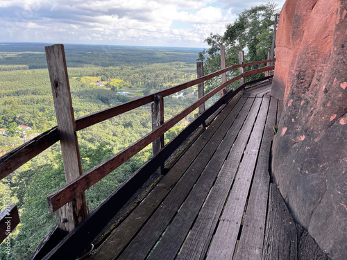 Wooden walkway around the steep sandstone cliff at Wat Phu Tok temple. Scenic mountain path trail with a wooden fence overlooking the green tropical forest and cloudy sky in Thailand