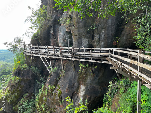 Wooden walkway around the steep sandstone cliff at Wat Phu Tok temple. Scenic mountain path trail with a wooden fence overlooking the green tropical forest and cloudy sky in Thailand