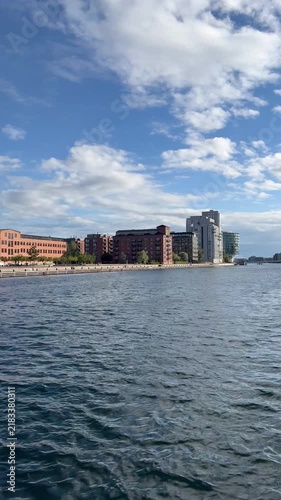 Scenic view of Islands Brygge waterfront in Copenhagen, Denmark from a boat navigating the calm waters of the Copenhagen Harbour on a bright, partly cloudy day - stock video