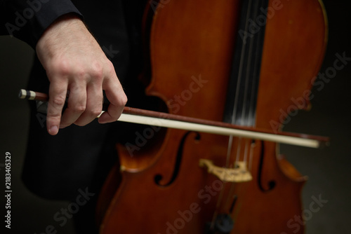 Caucasian young adult cellist hand holding bow playing acoustic cello in orchestral concert setting. Visual suitable for classical music education and cultural events promotion