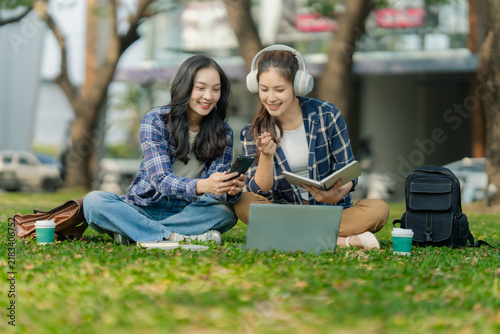 Two Asian female college students studying together and reading book on grass lawn in university campus.
