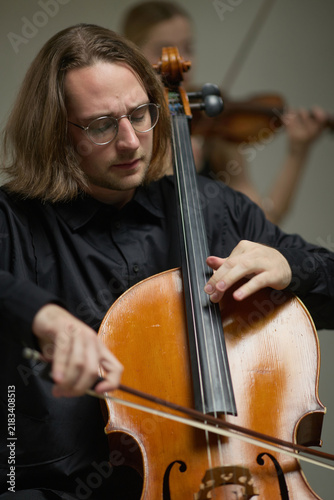 Vertical image of young adult Caucasian man playing cello during classical music orchestra rehearsal. Image suitable for arts education, concert advertising, symphonic performance promo