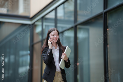 Confident Asian businesswoman using a smartphone and carrying a laptop in a modern office, representing digital communication, technology, and freelance career.