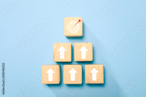 Wooden blocks with upward arrows lead to a target and arrow, symbolizing goal achievement, growth, strategy, success and business progress on a blue background with ample copy space.