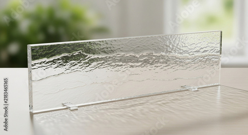 A textured glass panel stands upright on a wooden table inside a brightly lit room featuring a blurred green plant visible near the window during daylight hours for architectural interior design