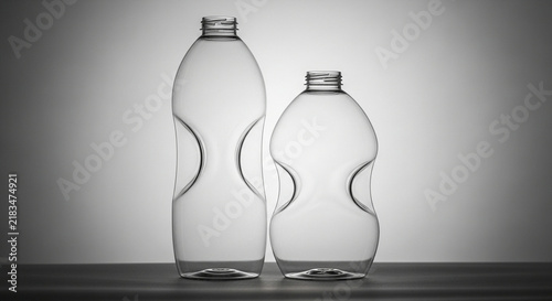 Two empty transparent plastic containers sit upright upon a dark horizontal wooden surface while dramatic overhead illumination highlights their unique curved silhouettes against a gradient wall