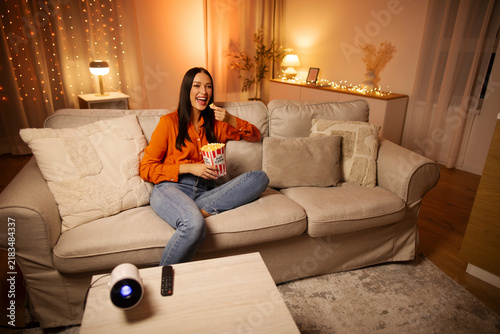 Young woman spending fun movie night at home, holding box of popcorn, sitting on comfy sofa in warmly lit living room, free space