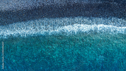 Top down aerial view with abstract background of blue turquoise sea waves crashing on a black pebble beach with white foam on a dark volcanic stone shore