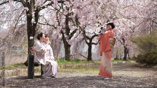 A kimono-clad person stands holding an infant beside two seated people under dense cherry blossom branches.
