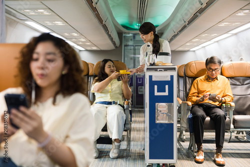 Photography Flight attendant serving drinks from a trolley in an airplane cabin as passengers sit and use smartphones; economy seating and aisle view create a realistic inflight travel scene