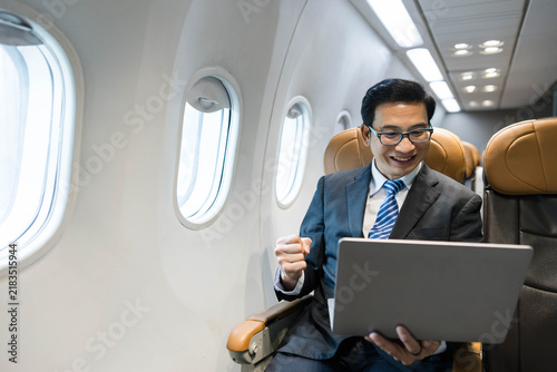 Foto Smiling businessman on airplane using laptop, fist pumped in celebration, working during flight with inflight WiFi, modern business travel productivity in aircraft cabin