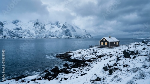 Small cabin with glowing windows by the sea, snowy mountain landscape under a moody overcast sky.