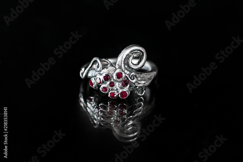 A silver ring with intricate designs and red gemstones, reflecting on a black surface. The ring showcases elegant craftsmanship and a luxurious appearance.