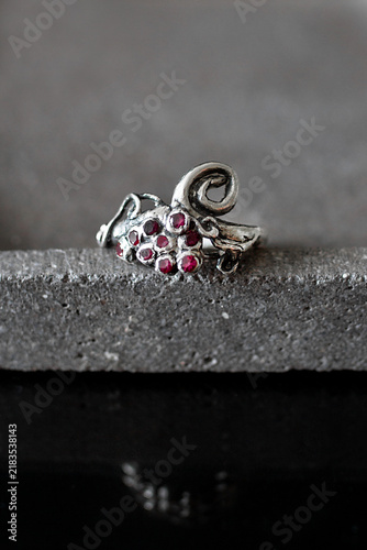 A silver ring designed with a grape cluster motif, featuring small red gemstones. The ring is placed on a textured gray surface, highlighting its intricate details.