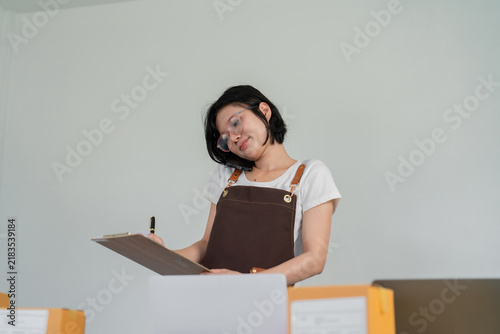 Small business owner holding phone to ear while writing on clipboard, standing at desk with laptop and shipping boxes, multitasking customer calls and orders