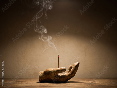 Ceramic hand incense burner with burning incense stick and delicate rising smoke on rustic wooden table against warm dark background