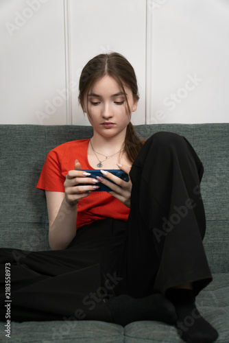 Obraz na płótnie Teenager girl is sitting comfortably on a gray sofa, engrossed in her smartphone, possibly browsing social media, playing games, or texting